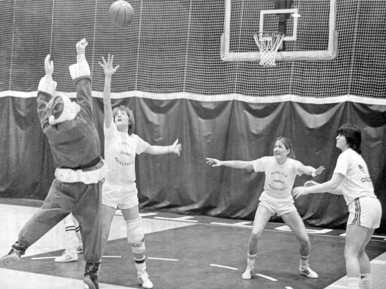 Santa Claus takes a jump shot while being guarded by one female basketball player while two other players defend and watch.